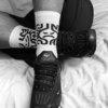 A high-contrast black-and-white photo of a person's legs wearing white SNIFFAIR CUM CUNT socks with black graphic patterns. The socks are paired with black Nike Air Max Plus sneakers on a white wrinkled bedsheet.