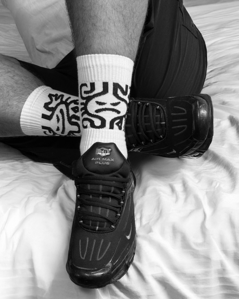 A high-contrast black-and-white photo of a person's legs wearing white SNIFFAIR CUM CUNT socks with black graphic patterns. The socks are paired with black Nike Air Max Plus sneakers on a white wrinkled bedsheet.